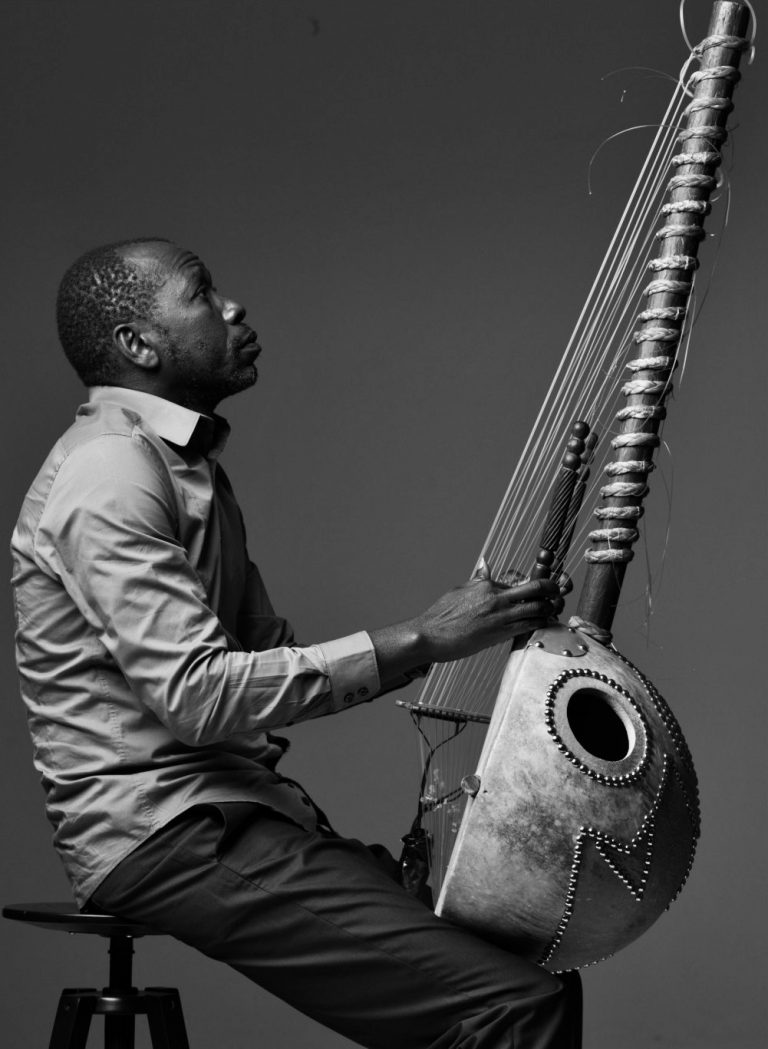 African man seated on a stool with his instrument, the kora, in his lap.